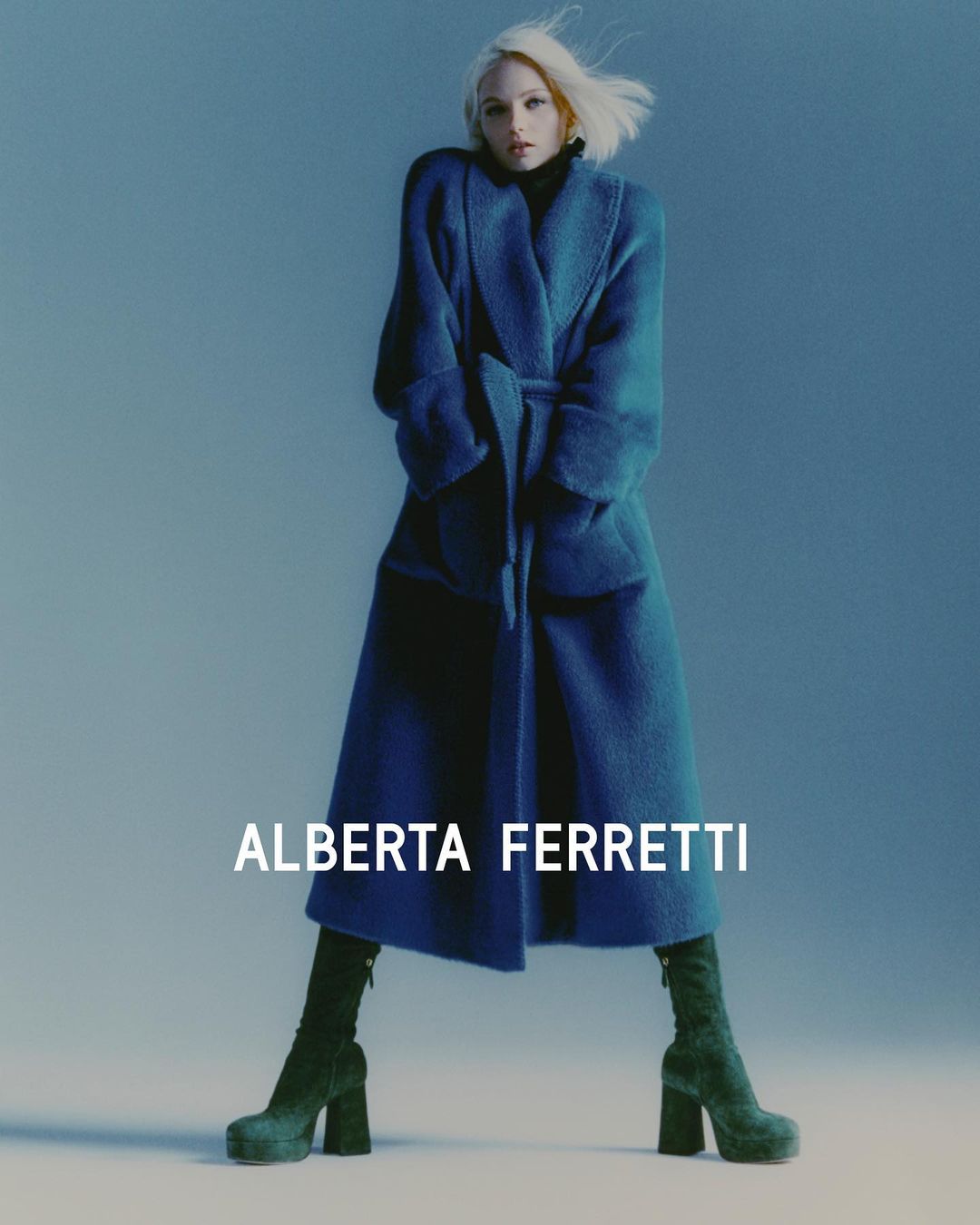 Fashion shot of a young adult woman wearing a blue coat with a tie belt and dark green platform boots against a blue studio backdrop.