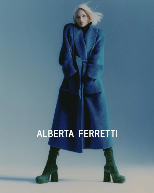Fashion shot of a young adult woman wearing a blue coat with a tie belt and dark green platform boots against a blue studio backdrop.
