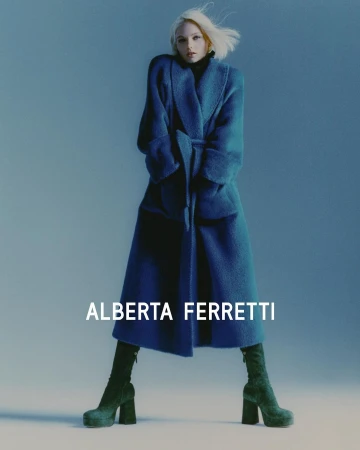 Fashion shot of a young adult woman wearing a blue coat with a tie belt and dark green platform boots against a blue studio backdrop.