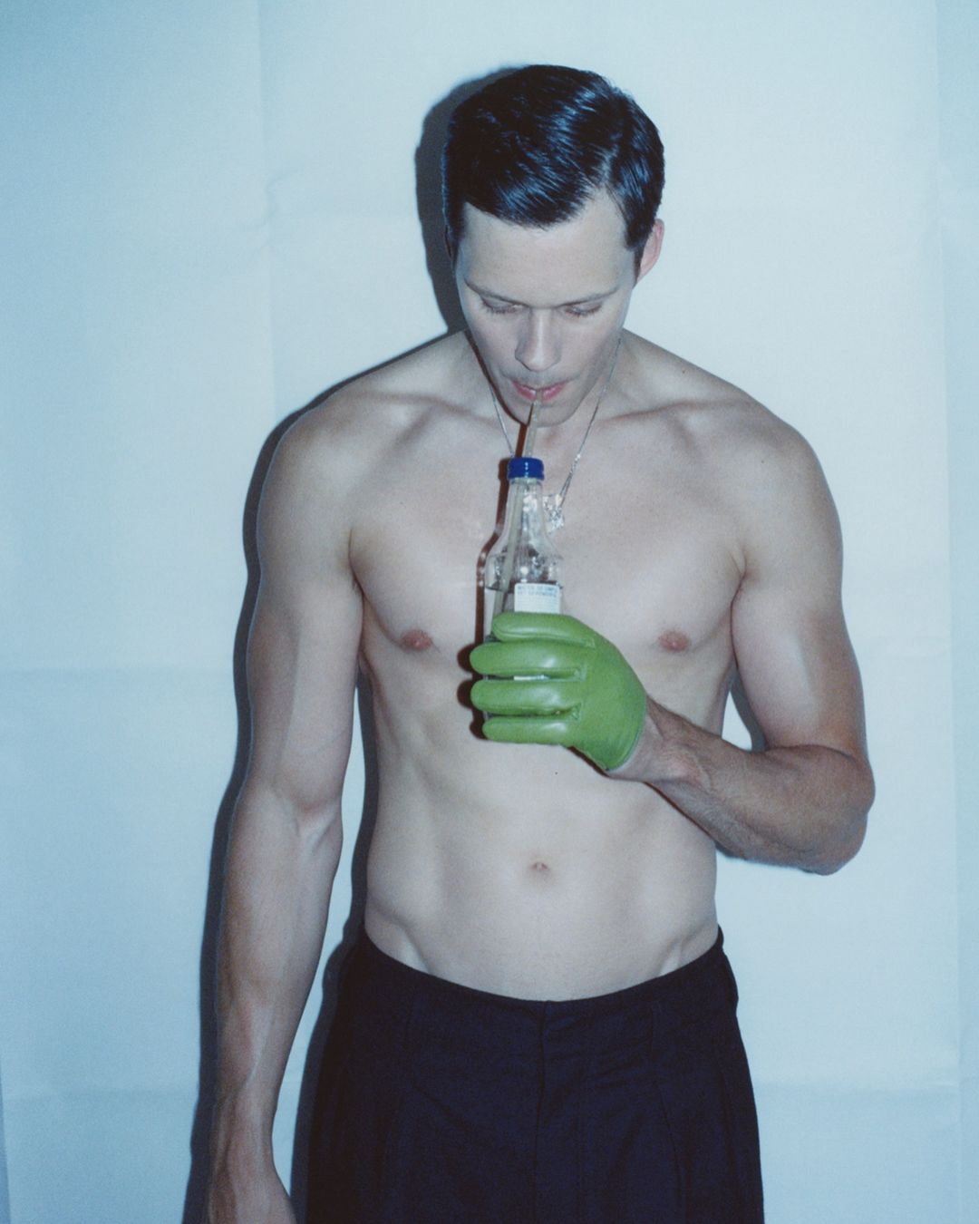 Shirtless man with dark hair drinks from a clear bottle with a straw and wears a green glove against a white background in studio lighting.
