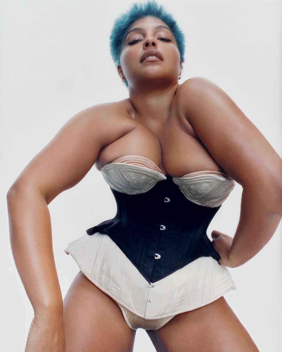 Eye-level studio shot of an adult female model with blue hair, posing in a black and beige corset against a white backdrop. 