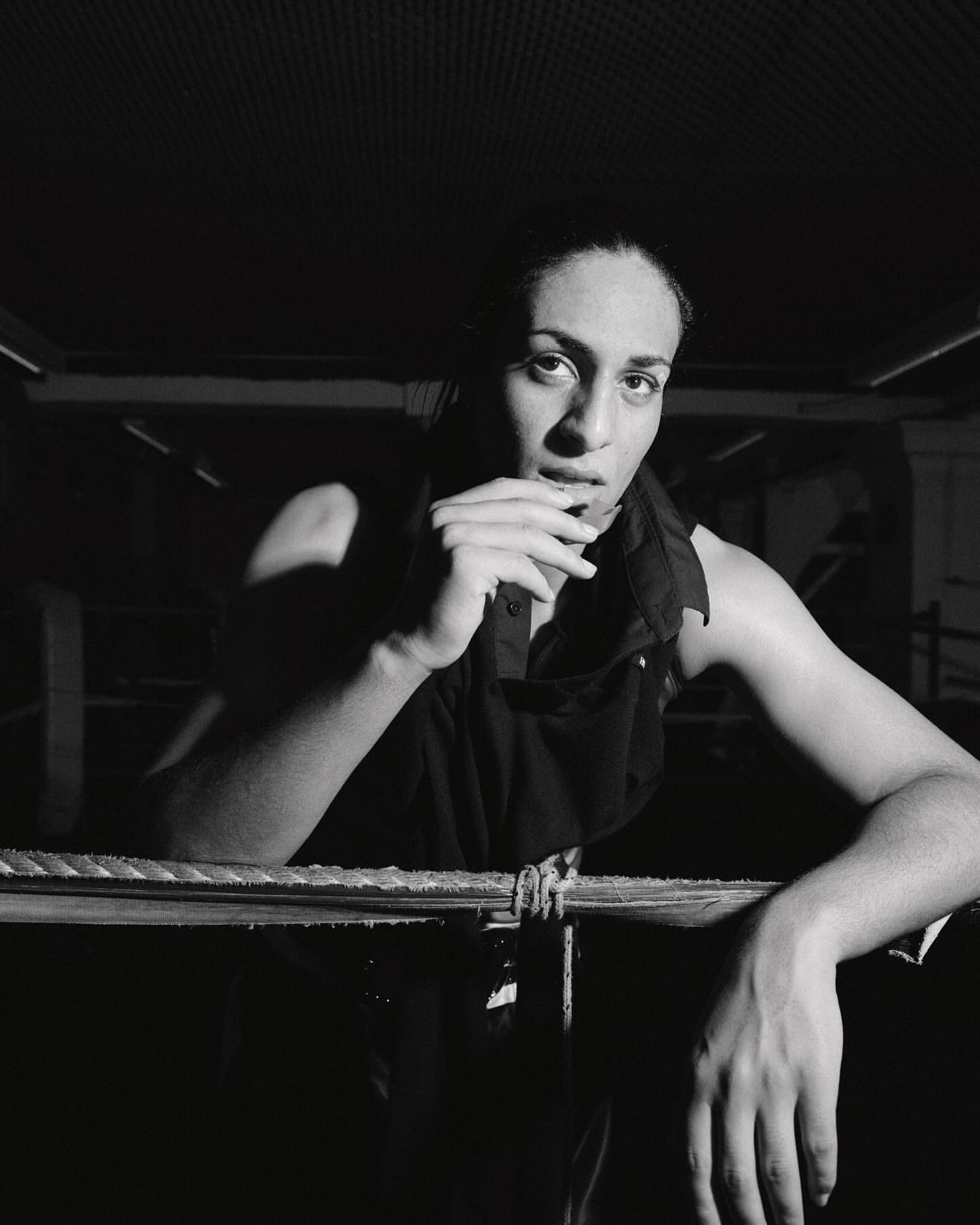 Monochrome portrait of a woman leaning on ropes, dressed in a dark sleeveless top, in a dimly lit indoor environment, creating an intense mood.
