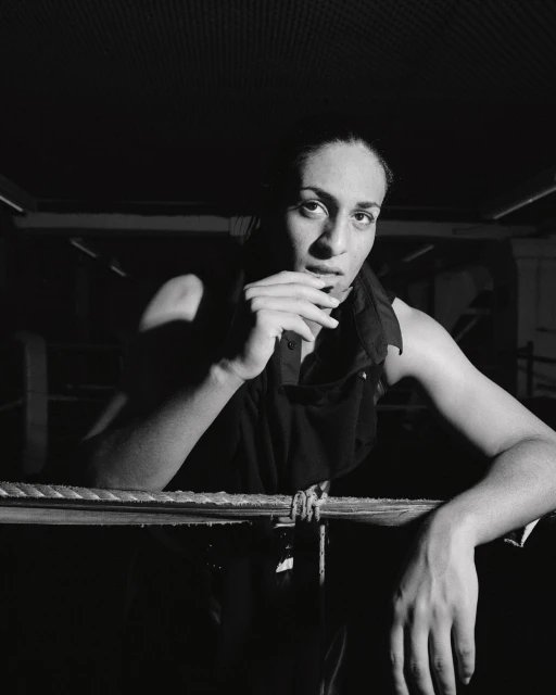 Monochrome portrait of a woman leaning on ropes, dressed in a dark sleeveless top, in a dimly lit indoor environment, creating an intense mood.