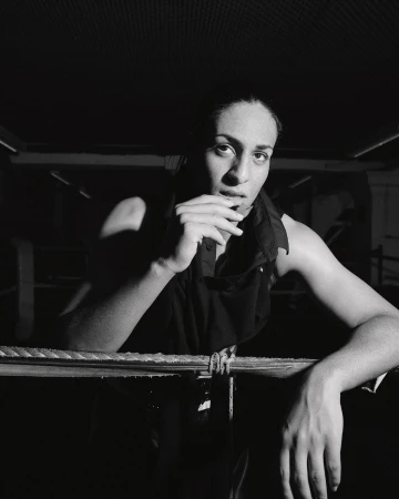 Monochrome portrait of a woman leaning on ropes, dressed in a dark sleeveless top, in a dimly lit indoor environment, creating an intense mood.