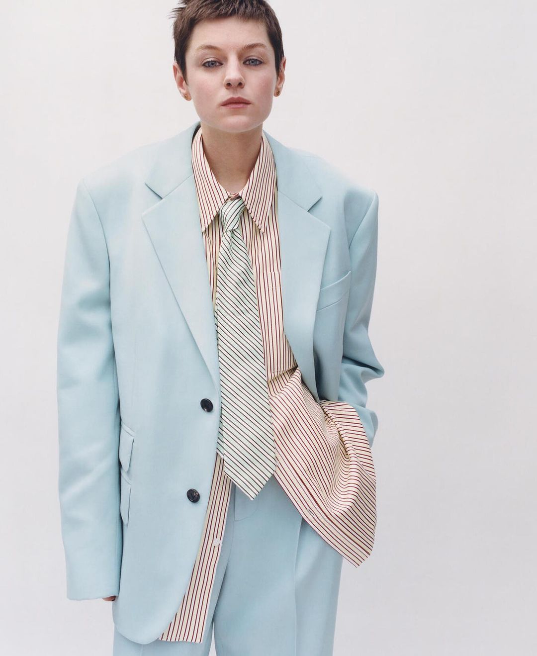 Woman in a light blue suit with a striped shirt and tie, posing in a studio against a white backdrop. Fashion editorial.