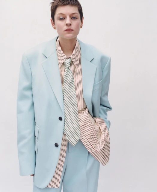 Woman in a light blue suit with a striped shirt and tie, posing in a studio against a white backdrop. Fashion editorial.