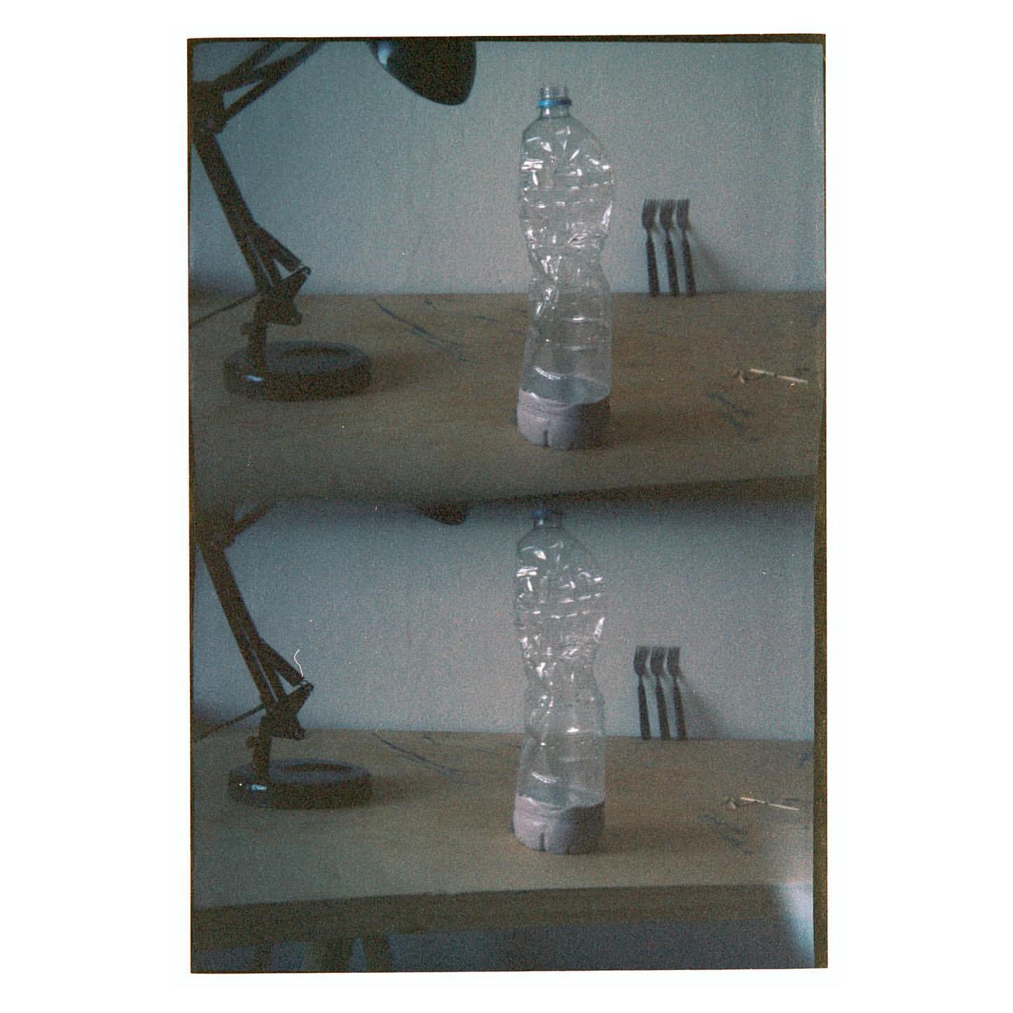 Still life featuring a distorted plastic water bottle on a wooden table, illuminated by a black desk lamp. Three metal objects in background.