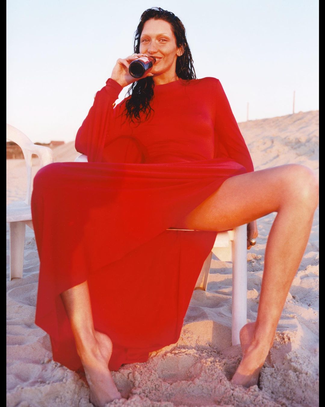 Elegant woman in a red dress sitting on a white plastic chair on a sandy beach, drinking from a can. Sky is light blue.