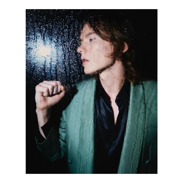 Portrait of a fair-skinned adult male with long hair, wearing a teal blazer and black shirt, posing in front of a window with raindrops.