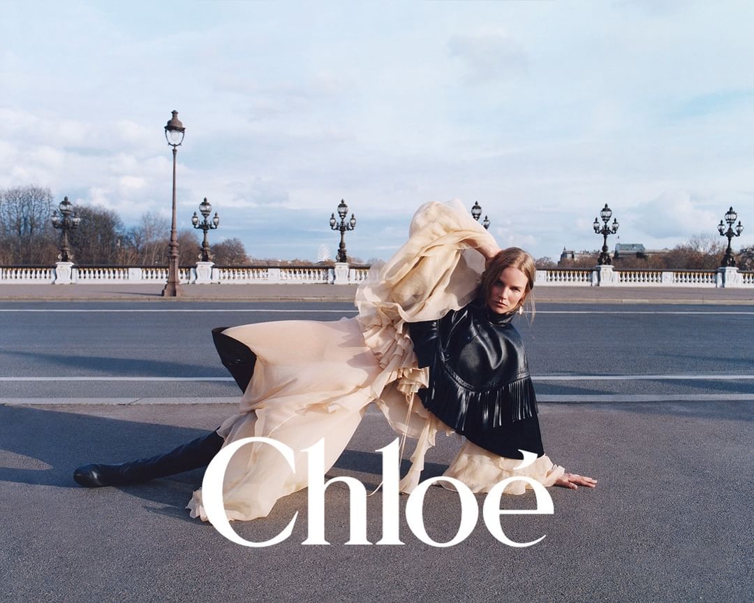 Fashion editorial shot of a blonde female model in draped beige dress with a black leather top on a bridge with lampposts.