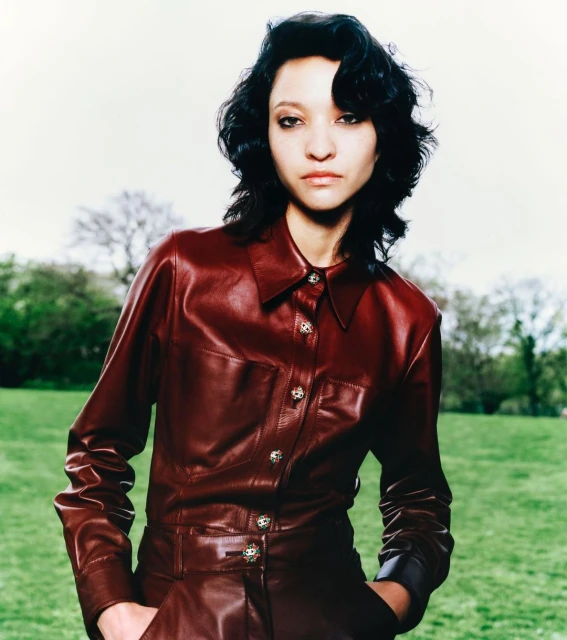 A portrait of a woman with dark wavy hair in a red leather collared button-up shirt posing in a grassy field with a backdrop of trees.