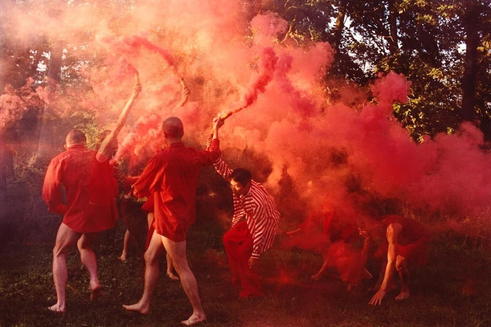 A group of people outdoors surrounded by red smoke, some holding smoke flares in the air, with trees and grass in the background.