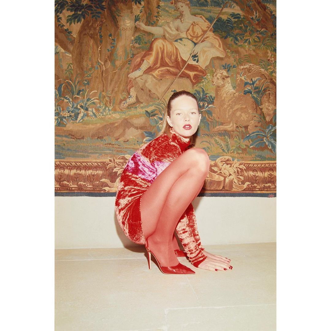 Young woman in red dress and heels poses in a crouch with a tapestry backdrop; fashion, beauty, and indoor studio aesthetic.