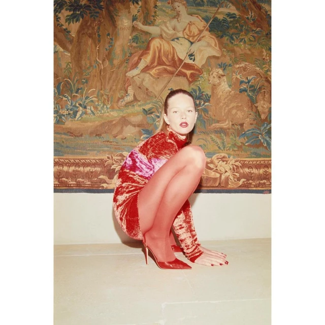Young woman in red dress and heels poses in a crouch with a tapestry backdrop; fashion, beauty, and indoor studio aesthetic.