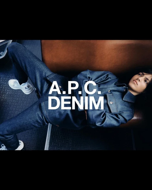 Brunette woman in denim jacket and jeans, lying against a brown backrest with white sneakers in an indoor setting with A.P.C. Denim text.