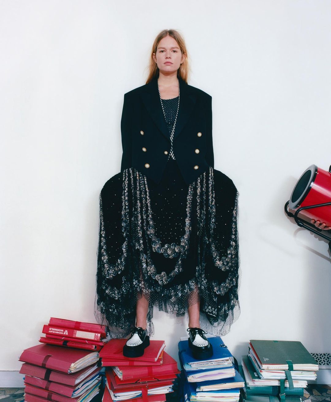 Blonde female fashion model stands on books against a white wall, wearing a black jacket, lace skirt, and platform shoes in a studio.