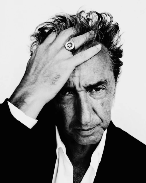 Black-and-white portrait of an adult male wearing a ring and suit coat, his hand touching his head, against a white backdrop.