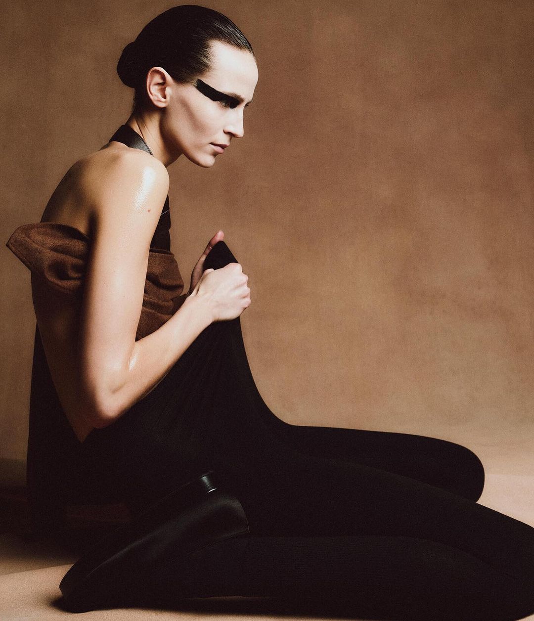 Elegant portrait of a slender adult woman in a black body suit, styled with brown draped fabric, dramatic makeup, and a minimalist studio.