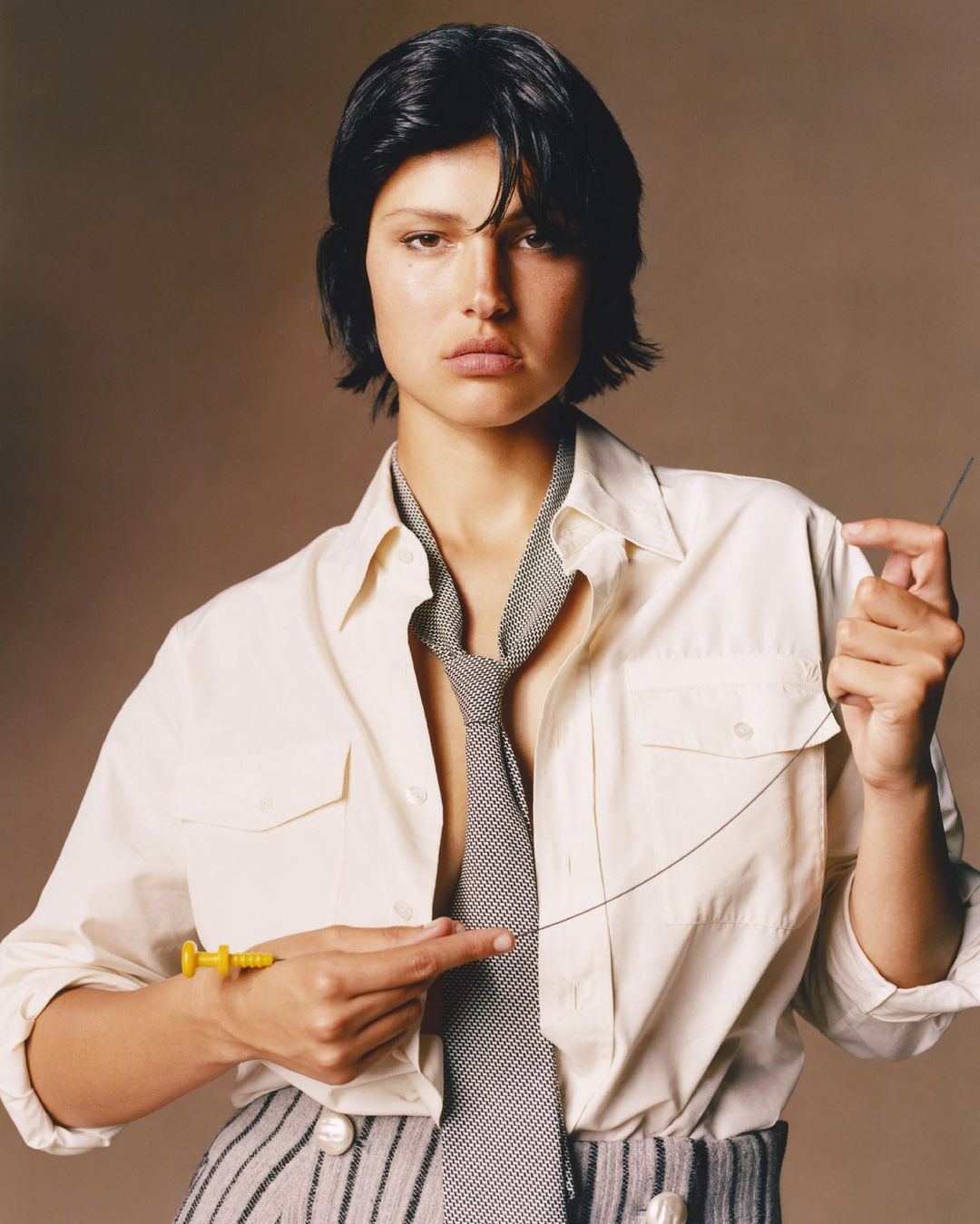 Portrait of a woman with dark hair in a white shirt and striped pants, holding a yellow syringe and a long needle in a studio.