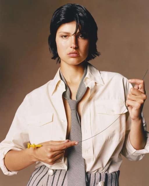 Portrait of a woman with dark hair in a white shirt and striped pants, holding a yellow syringe and a long needle in a studio.