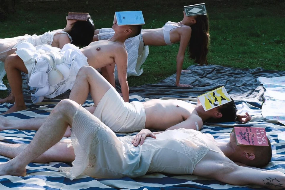 Group of people lie on a blue-and-white striped blanket in a grassy outdoor setting with books covering their faces.