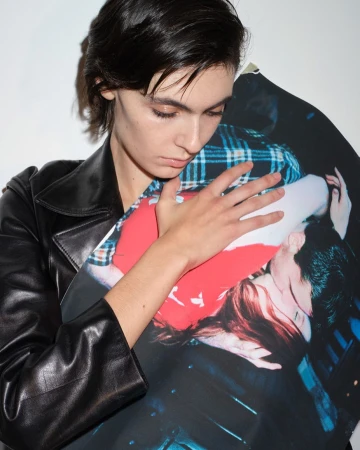 Adult female with dark hair holding a photograph of a couple in a close embrace, wearing a black leather jacket, against a white background.