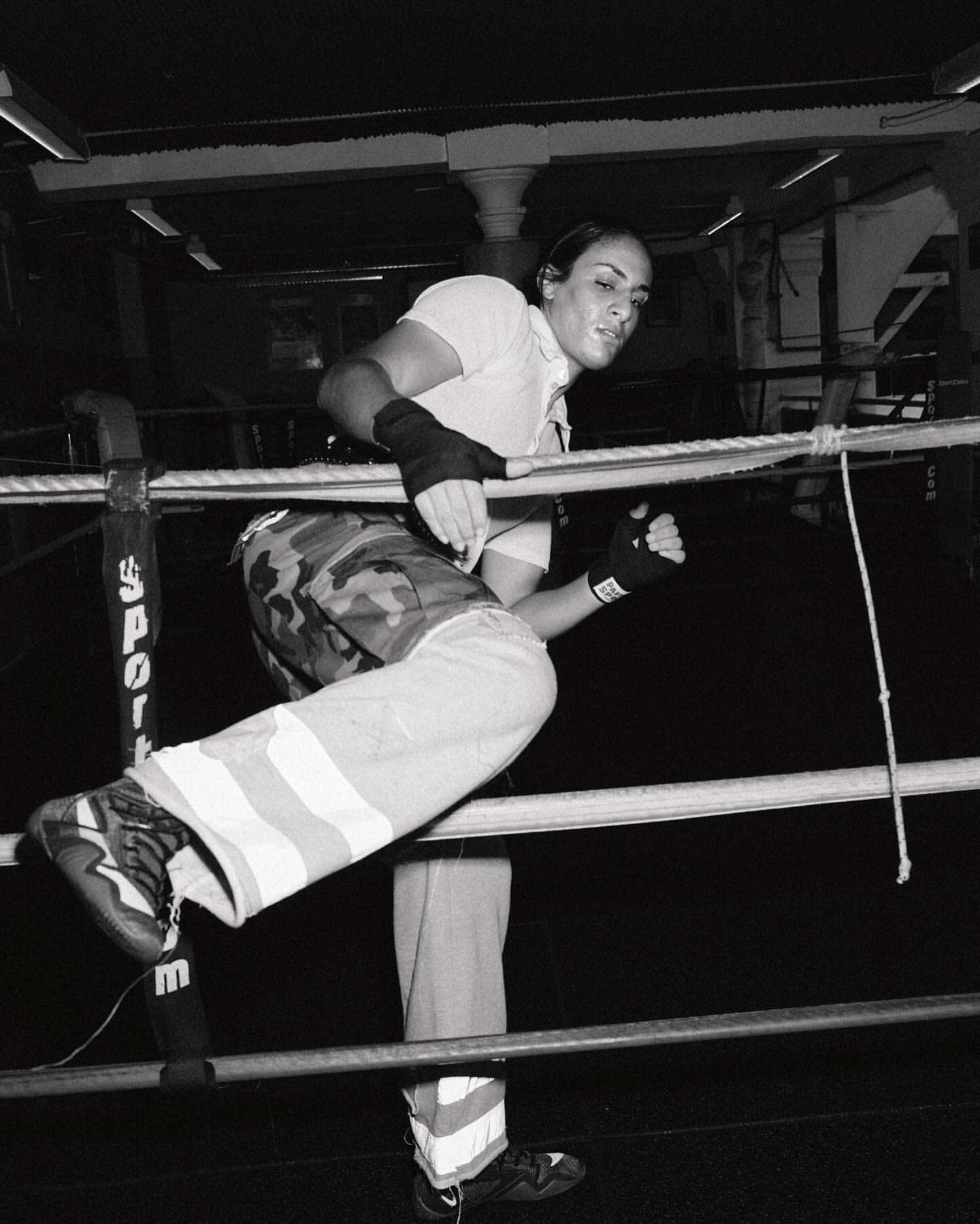 Female boxer in camo pants climbs through boxing ring ropes in black and white, preparing for a match, with a determined expression.