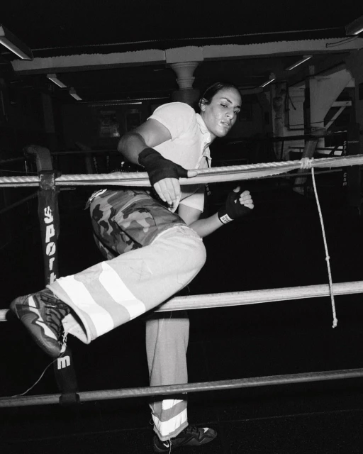 Female boxer in camo pants climbs through boxing ring ropes in black and white, preparing for a match, with a determined expression.