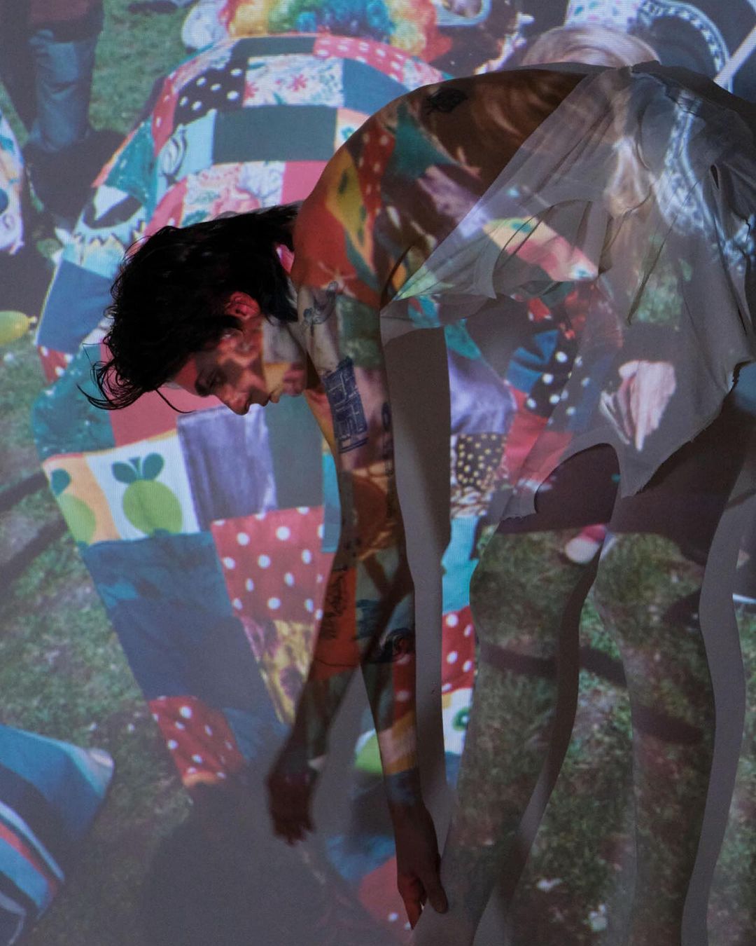 Adult bending forward with a colorful patchwork design projected over their body, casting a ghostly figure on the white wall.