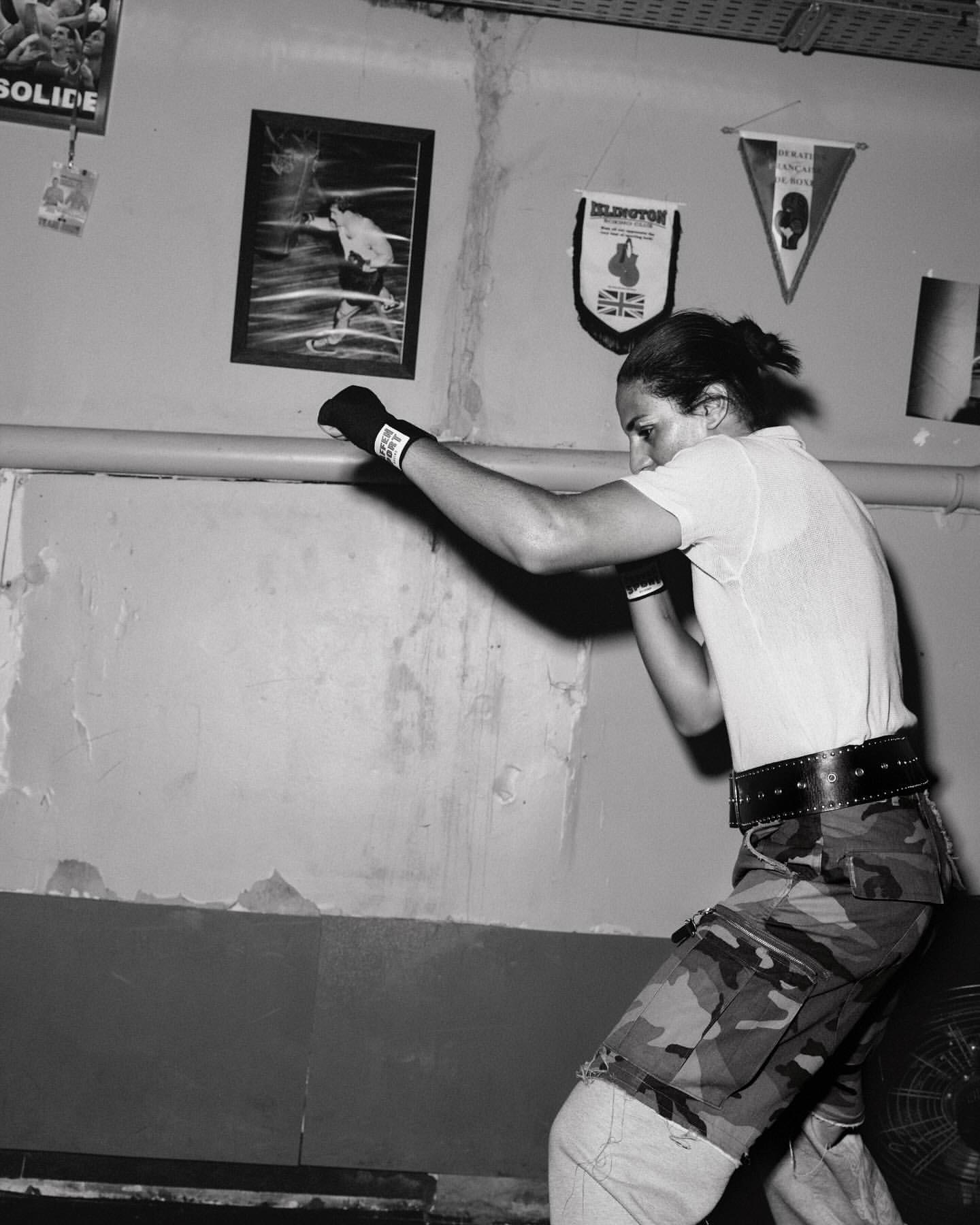 Monochrome image of a brunette male boxer in mid-punch in an indoor boxing gym, featuring faded posters and flags in the background.
