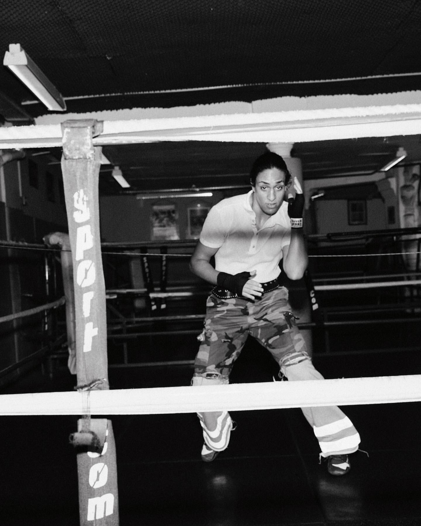Monochrome image of a female boxer standing in a boxing ring, wearing camouflage pants and boxing wraps, looking focused and ready.
