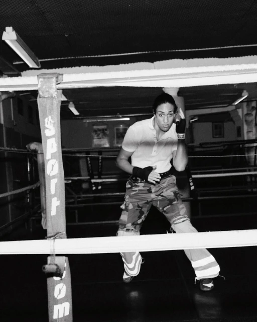 Monochrome image of a female boxer standing in a boxing ring, wearing camouflage pants and boxing wraps, looking focused and ready.