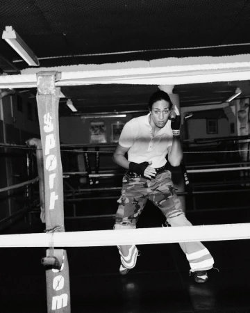 Monochrome image of a female boxer standing in a boxing ring, wearing camouflage pants and boxing wraps, looking focused and ready.