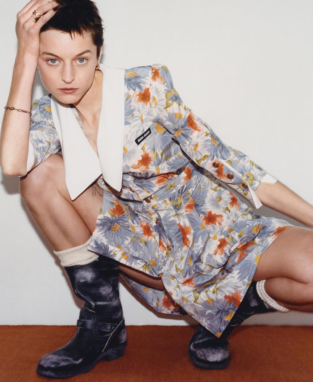 A woman in a floral dress and black boots poses against a white wall and brown floor, striking a confident and fashionable pose.