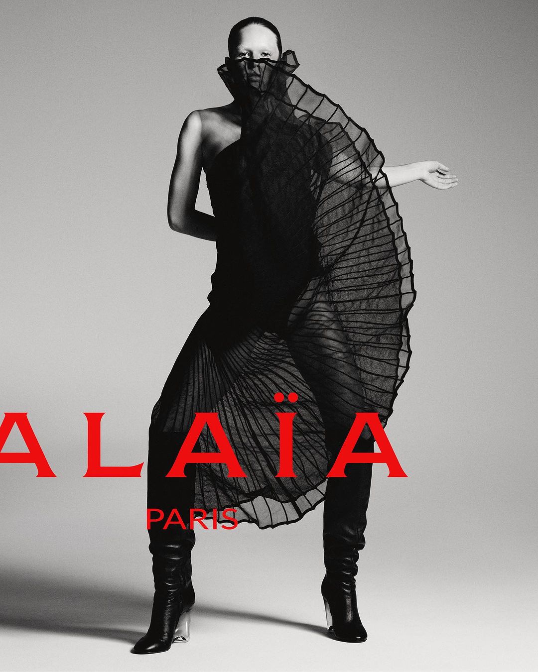 Fashion editorial shot of a single bald adult female posing in a black dress and black boots against a grey studio backdrop.