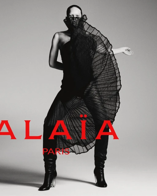 Fashion editorial shot of a single bald adult female posing in a black dress and black boots against a grey studio backdrop.