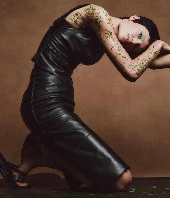 A woman with light skin poses while kneeling and arching her back. She is wearing a black dress and strappy heels against a tan background.