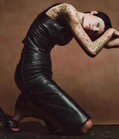 A woman with light skin poses while kneeling and arching her back. She is wearing a black dress and strappy heels against a tan background.