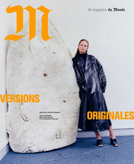 Fashion editorial cover shot: Adult woman poses in shiny coat next to white plaster object with magazine text. Studio lighting. 