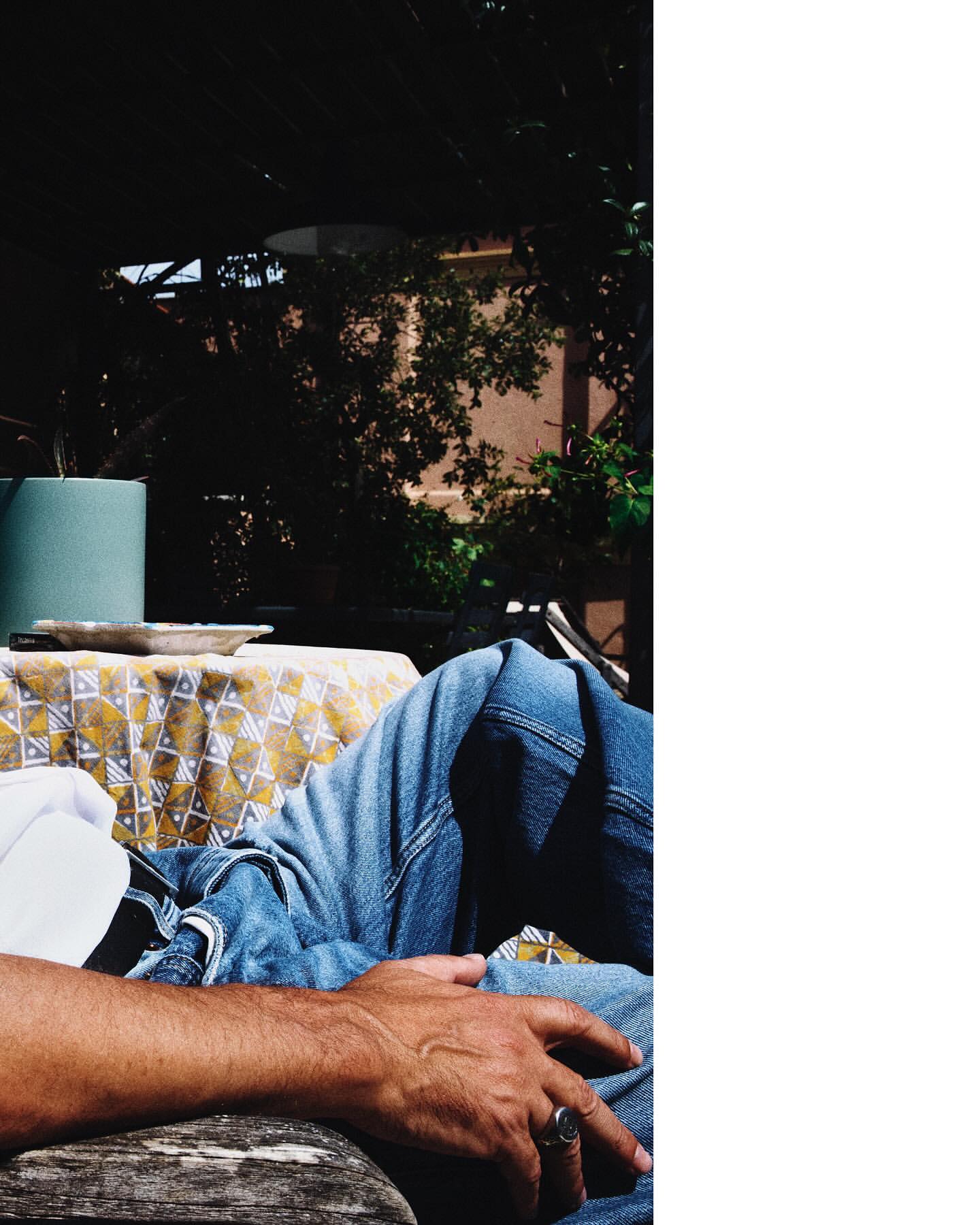 Person resting outdoors in blue jeans with yellow patterned tablecloth, surrounded by lush greenery, bathed in soft sunlight.