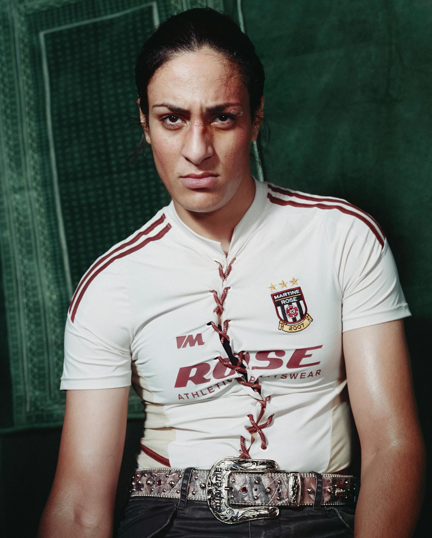 Woman with a determined expression, wearing a white sports jersey and an elaborate belt, poses against a textured green backdrop.