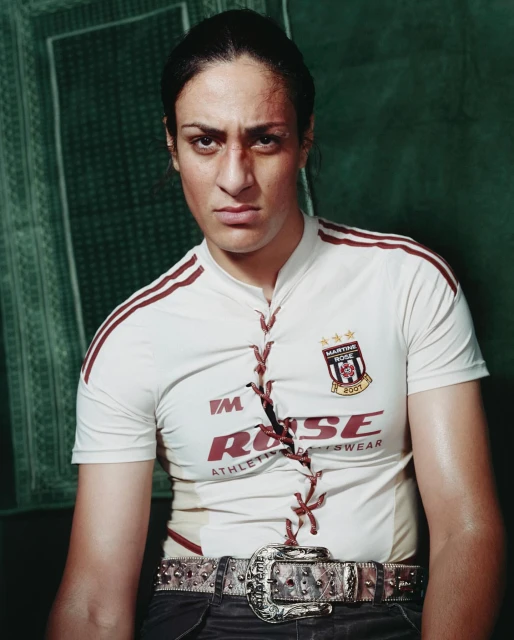 Woman with a determined expression, wearing a white sports jersey and an elaborate belt, poses against a textured green backdrop.