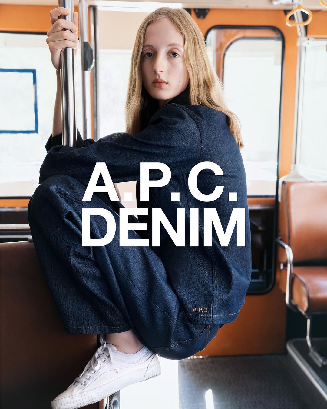 Young woman in denim outfit sitting inside a bus, posing for a fashion advertisement with 'A.P.C. DENIM' text overlay.