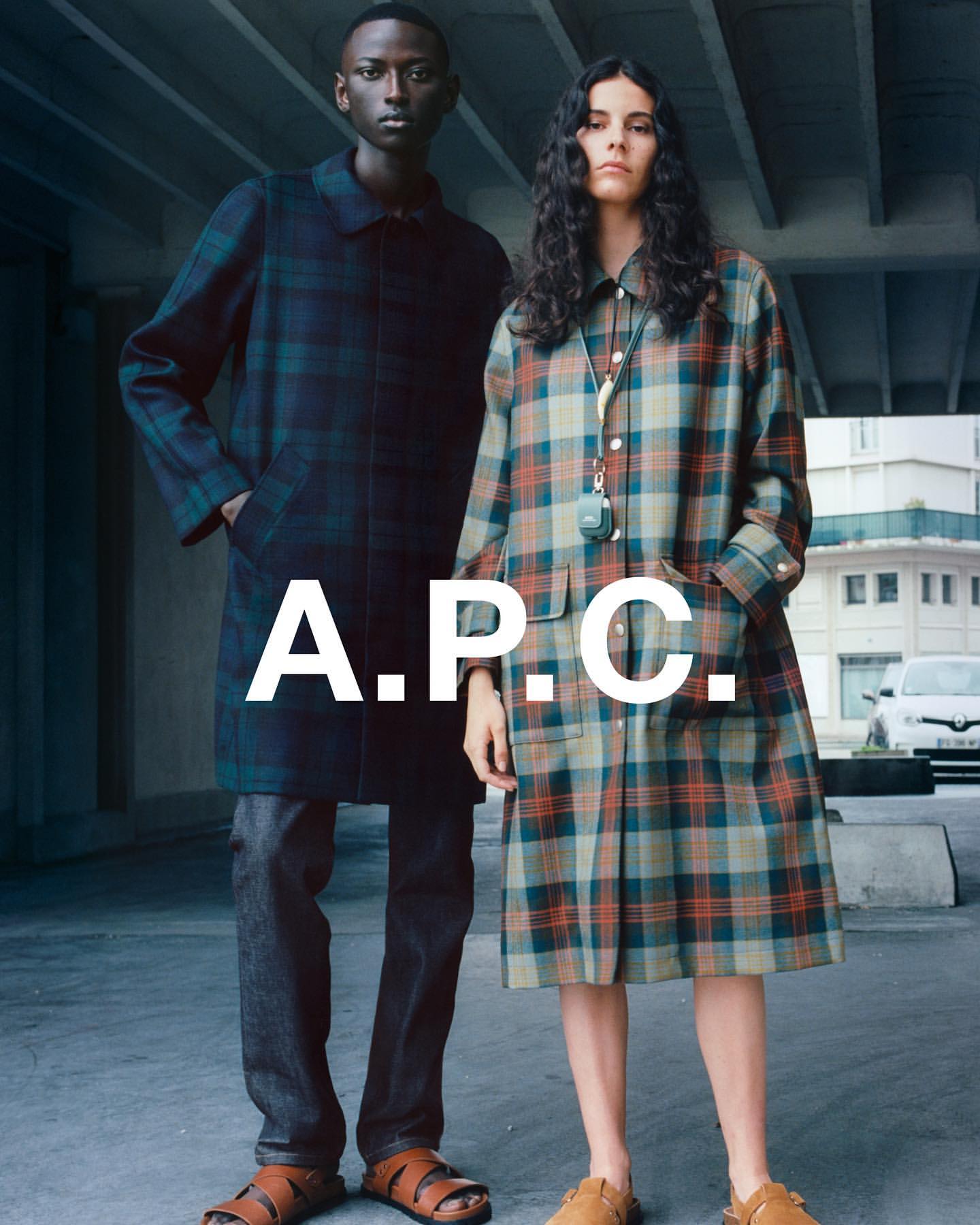 Two models in plaid coats stand against a concrete backdrop with a parked car; the A.P.C. logo is superimposed in the middle.