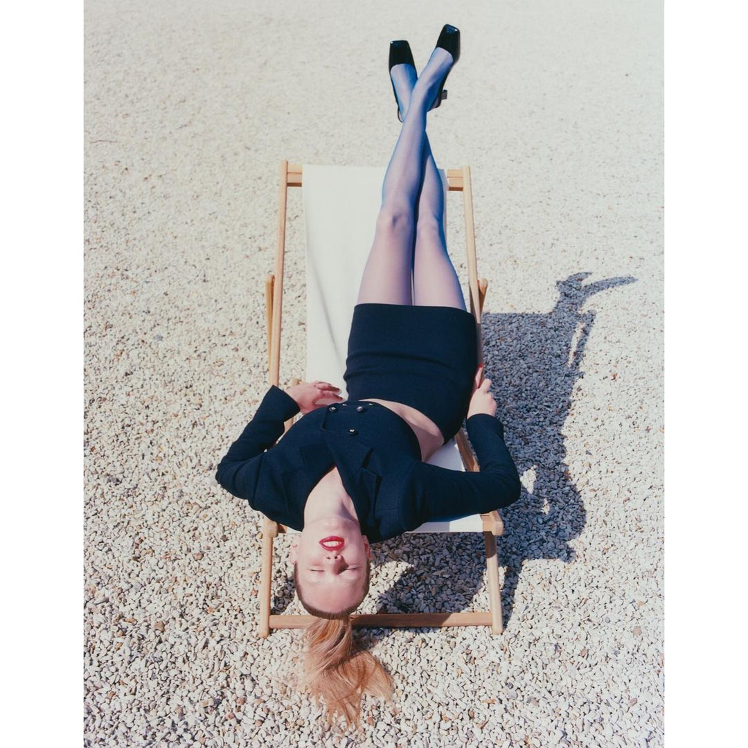 Woman in black skirt suit and hosiery lounging on a deck chair in the sun outside on a stone surface in outdoor setting.