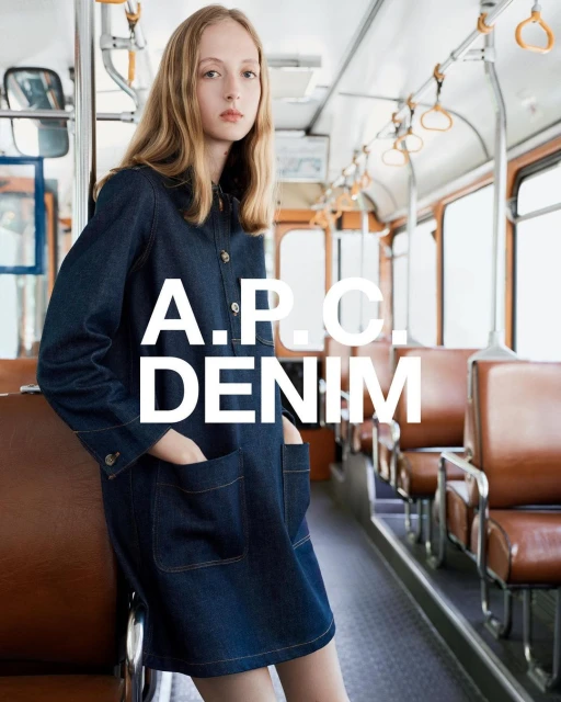 A blonde woman poses in an A.P.C. denim dress inside a vintage bus with tan leather seats. A.P.C. Denim logo displayed.