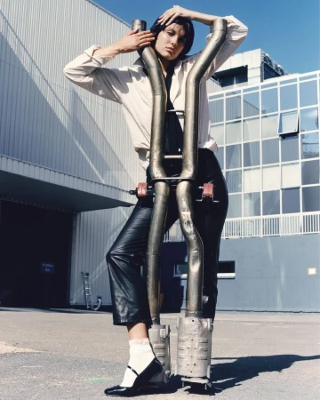 A woman poses with car exhaust pipes in front of a building, wearing a white blouse, black leather pants, and Mary Jane shoes with white socks.