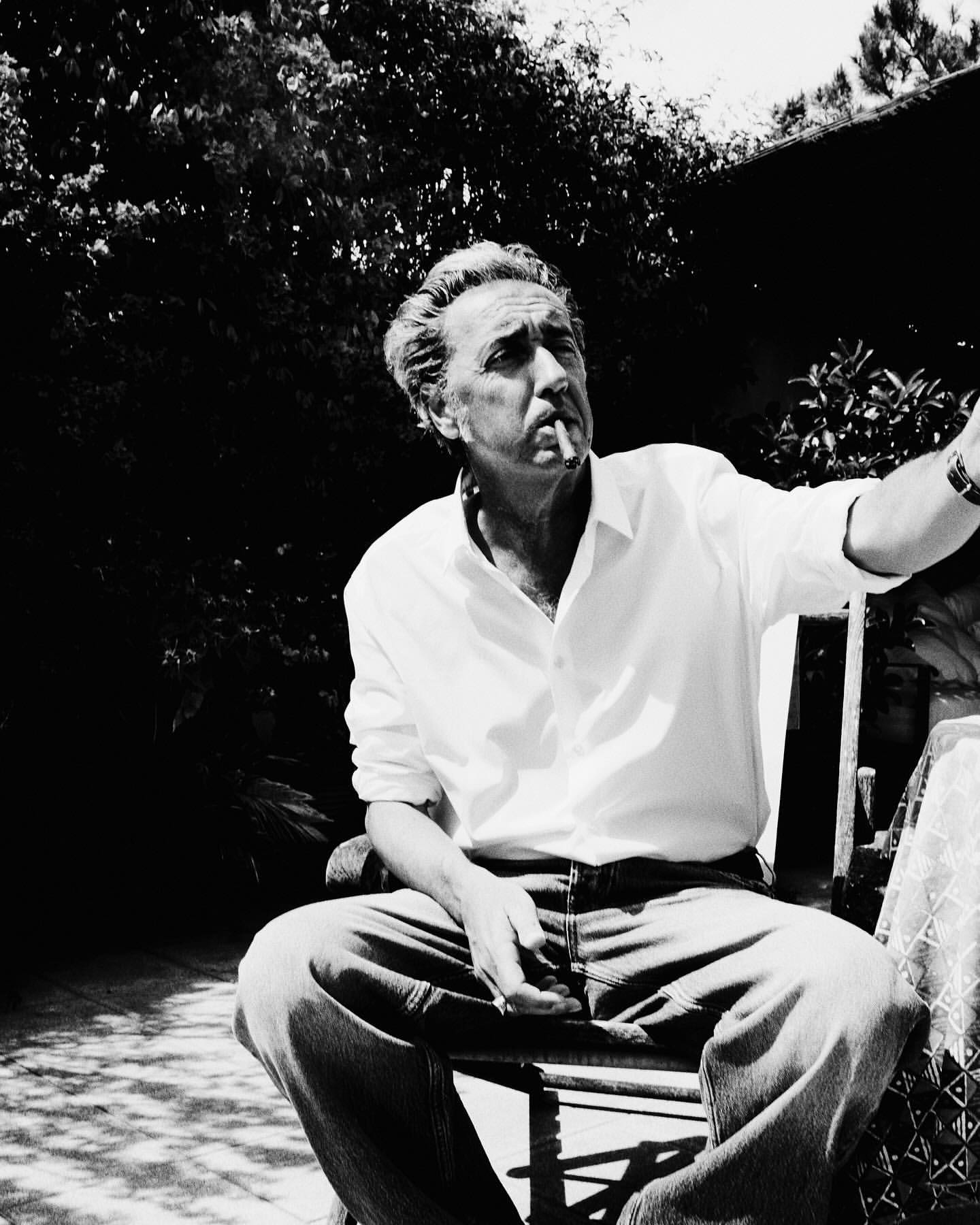 Black and white portrait of an adult man smoking outdoors. He's wearing a white shirt and jeans while sitting on a wooden chair.