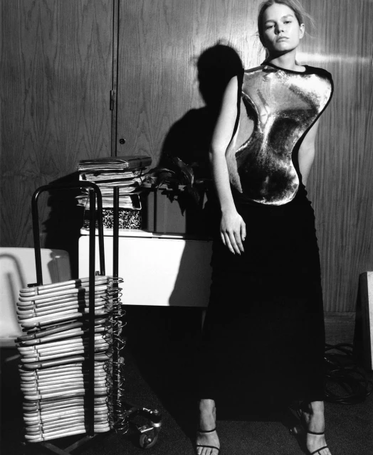 Monochrome portrait of a woman in a silver top and black skirt, indoors with a clothing rack and a wooden wall in the background.