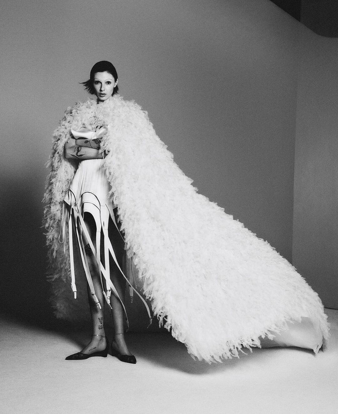 Black and white portrait of a fashionable adult woman in an off-white shaggy cloak, accessorized with leg garters in a studio setting.